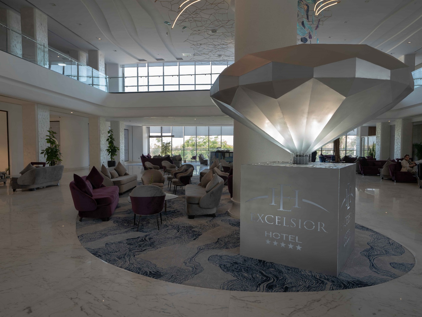 لابی هتل اکسلسیور با مبلمان رنگارنگ شامل صندلیهای بنفش و آبی، فرش زیبا و پنجرههای بزرگ که نمای بیرونی و فضای سبز را نمایش میدهد