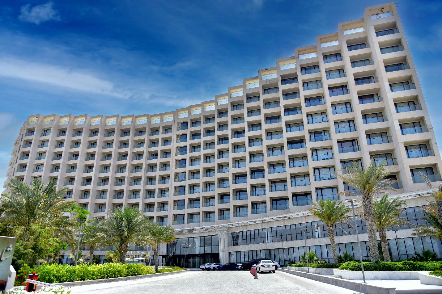 نمای خارجی اکسلسیور هتل مدرن با پنجرههای بزرگ، درختان نخل و فضای سبز، ورودی هتل و خودروهای پارک شده در مقابل